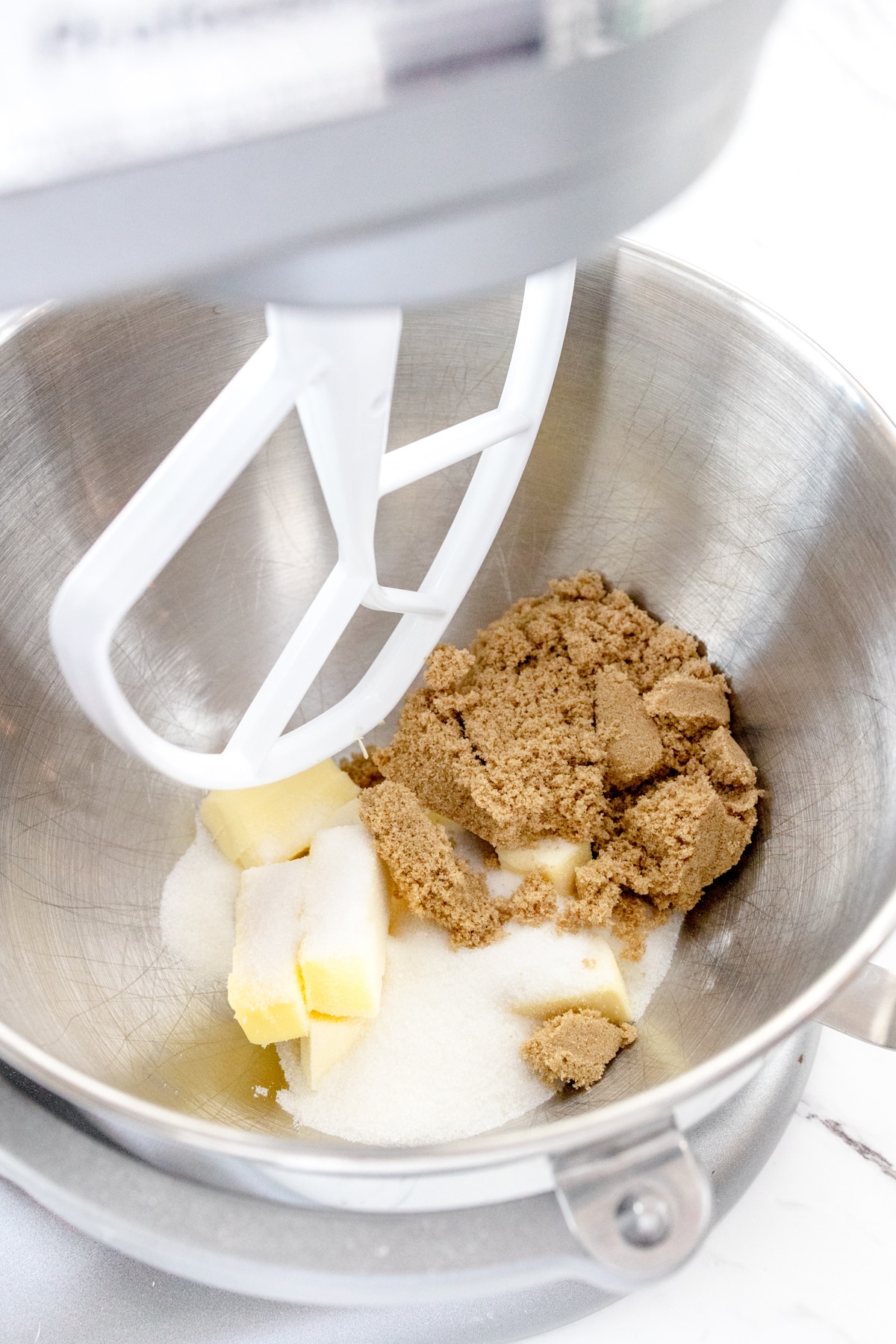 Close up of the bowl of a stand mixer with butter, granulated sugar, and brown sugar about to be creamed together with a paddle attachment.