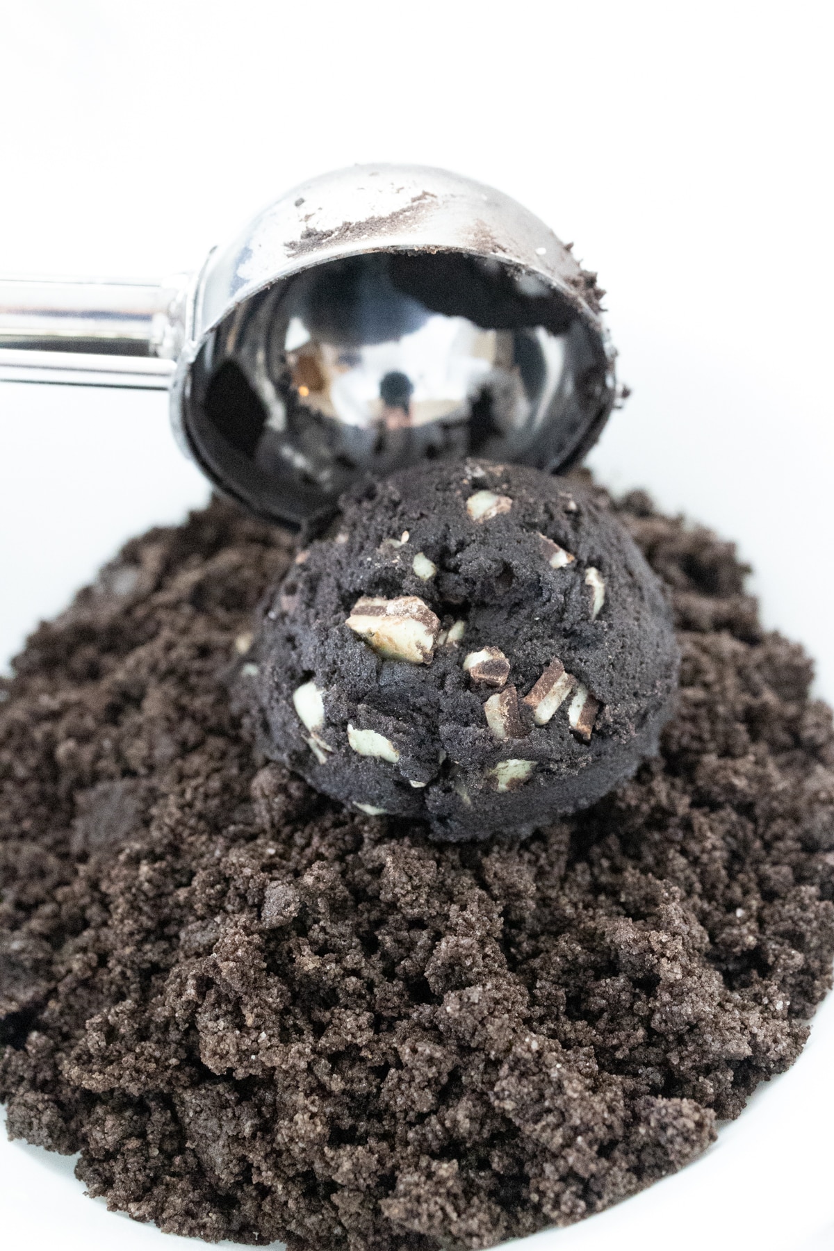 Close up of a cookie scoop placing a scoop of cookie dough into a bowl of oreo cookie crumbs.