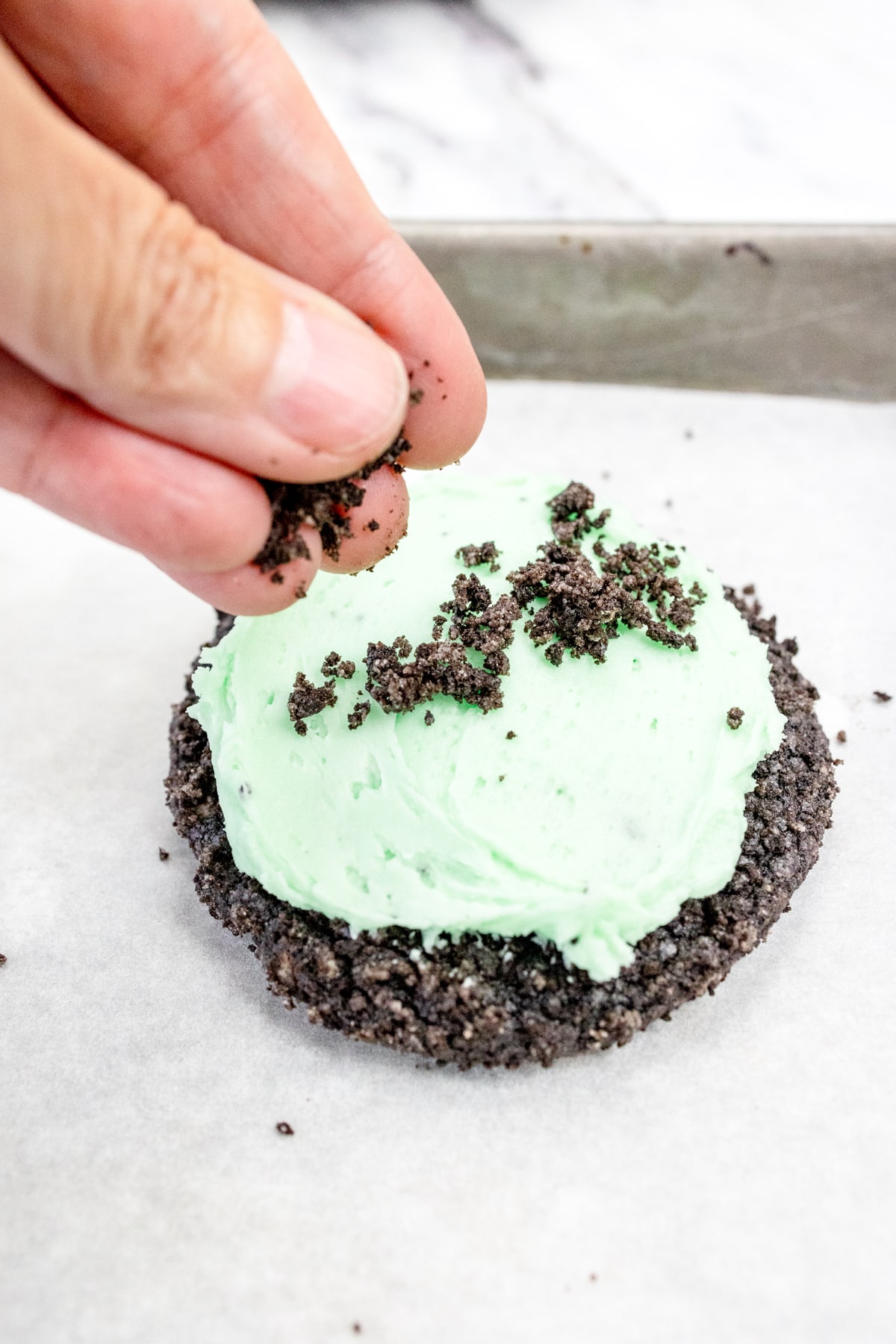 Close up of a hand sprinkling oreo crumbs on top of a frosted mint oreo cookie.