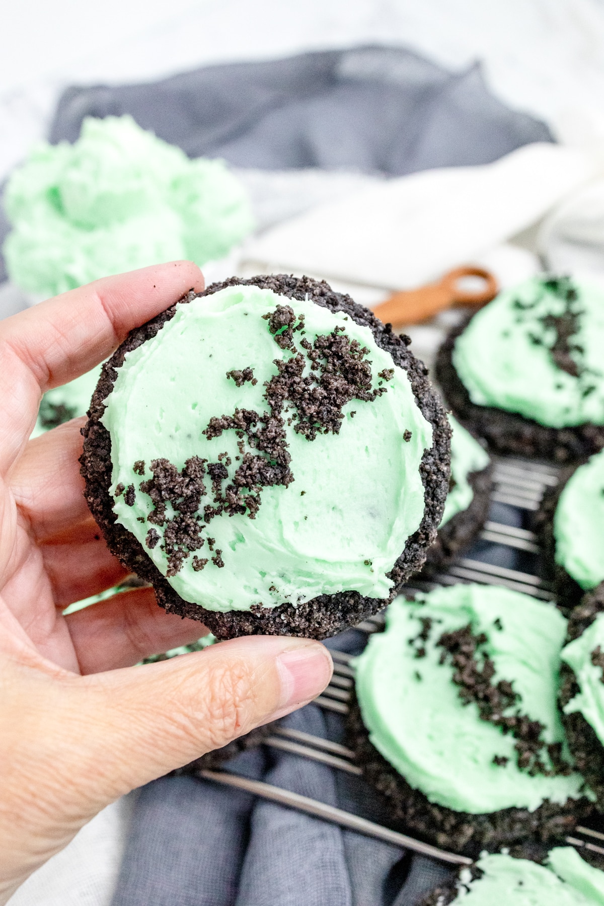 Close up of a hand holding a Frosted Mint Oreo Cookie above other cookies on a wire rack, with a bowl of frosting and a spoon full of oreo crumbs next to them.