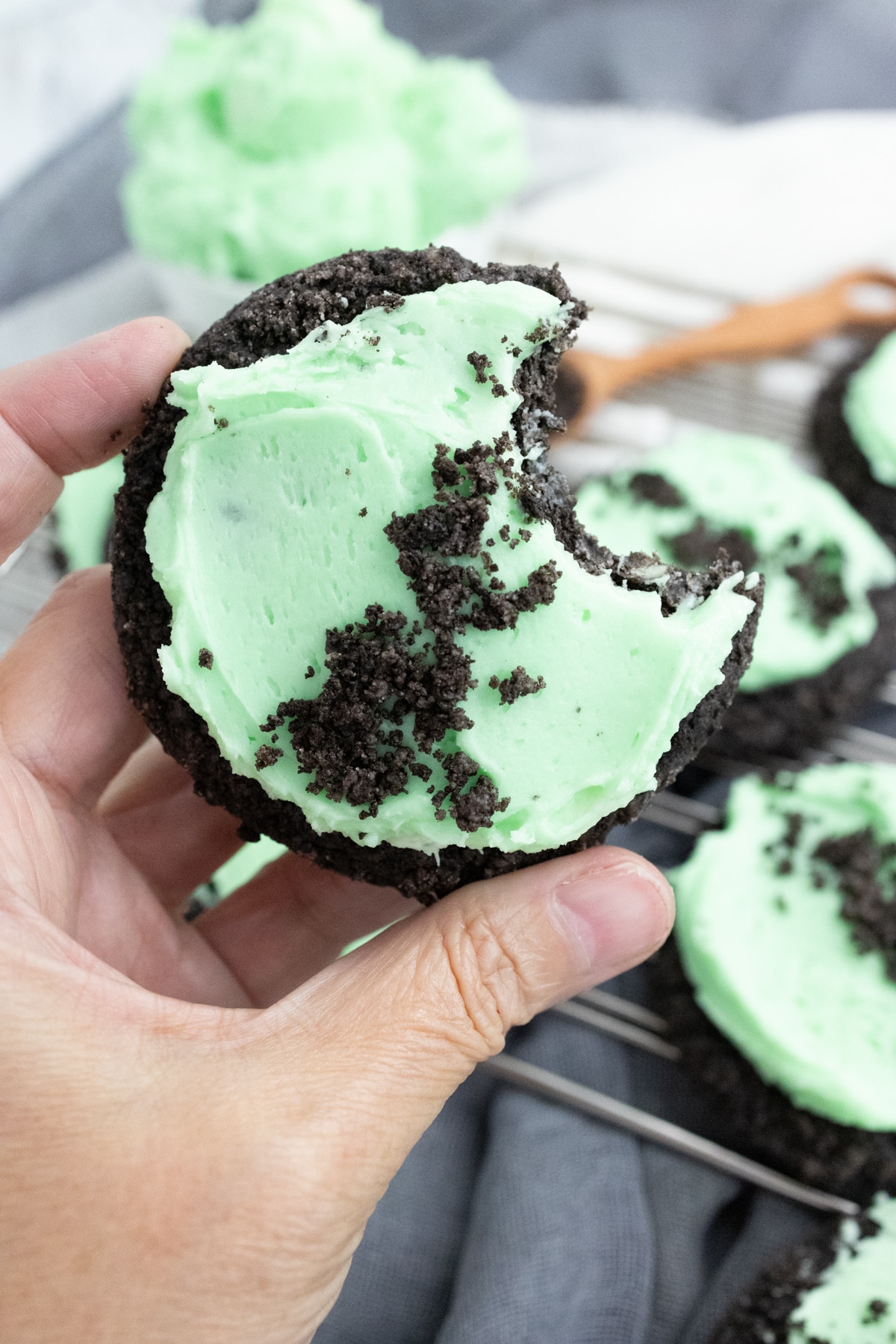 Close up of a hand holding a Frosted Mint Oreo Cookie with a bite taken out of it, above other cookies on a wire rack, with a bowl of frosting and a spoon full of oreo crumbs next to them.