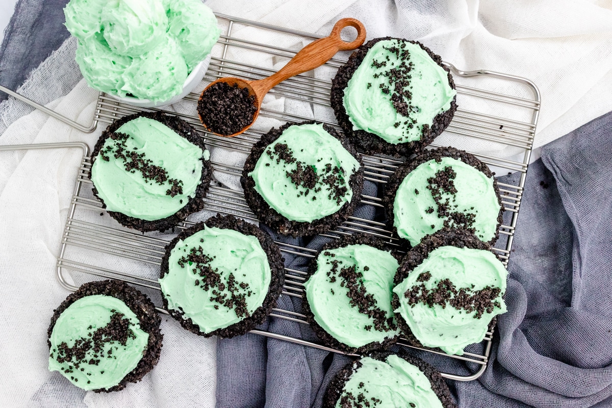 Top view of Frosted Mint Oreo Cookies on a wire rack, with a bowl of frosting and a spoon full of oreo crumbs next to them.
