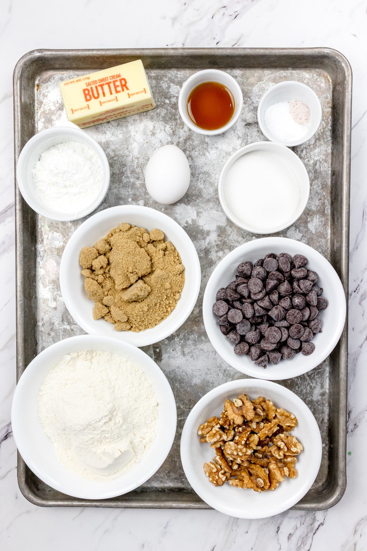 Top view of ingredients needed to make Chocolate Chip Walnut Cookies in small bowls on a baking tray.