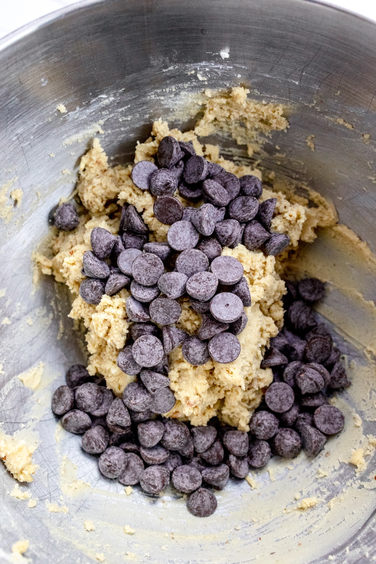 Close up of the bowl of a stand mixer with cookie dough in it with chocolate chips and walnuts that have been added on top to mix in.