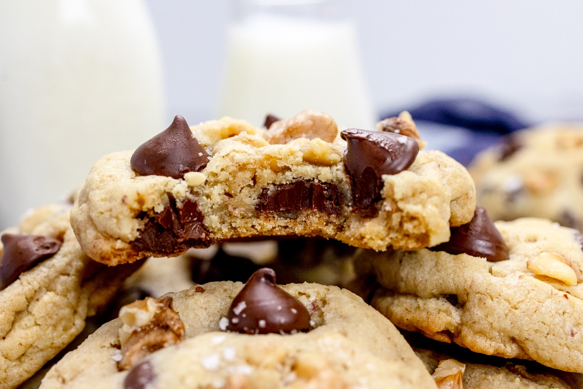 Close up view of Chocolate Chip Walnut Cookies on a white plate with glasses of milk beside it, and the cookie on top is sliced in half to reveal what's inside.