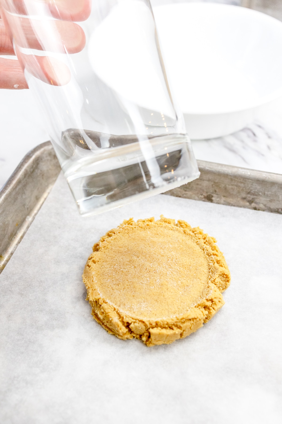 Close up of a drink glass pressing a scoop of cookie dough flat on a baking tray lined with parchment paper.