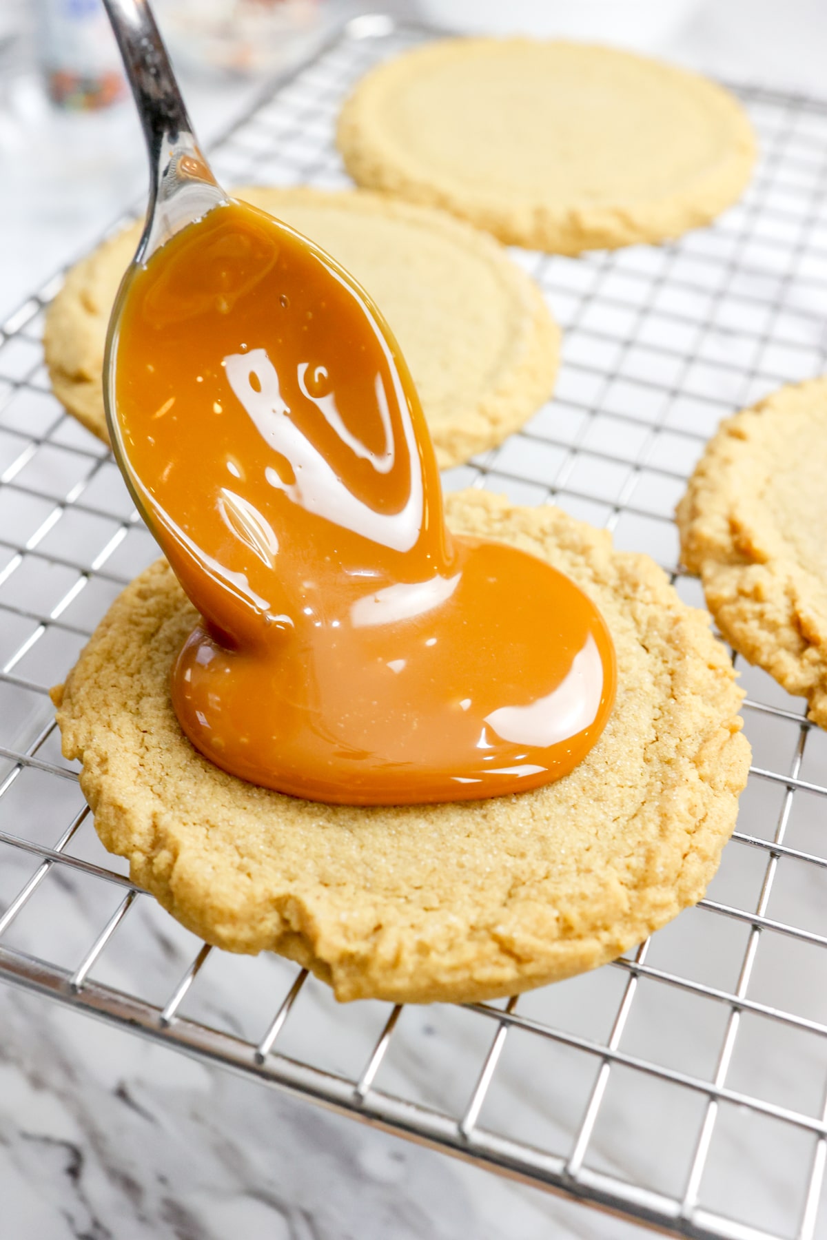 Close up of a cookie on a wire rack being topped with caramel using a spoon.