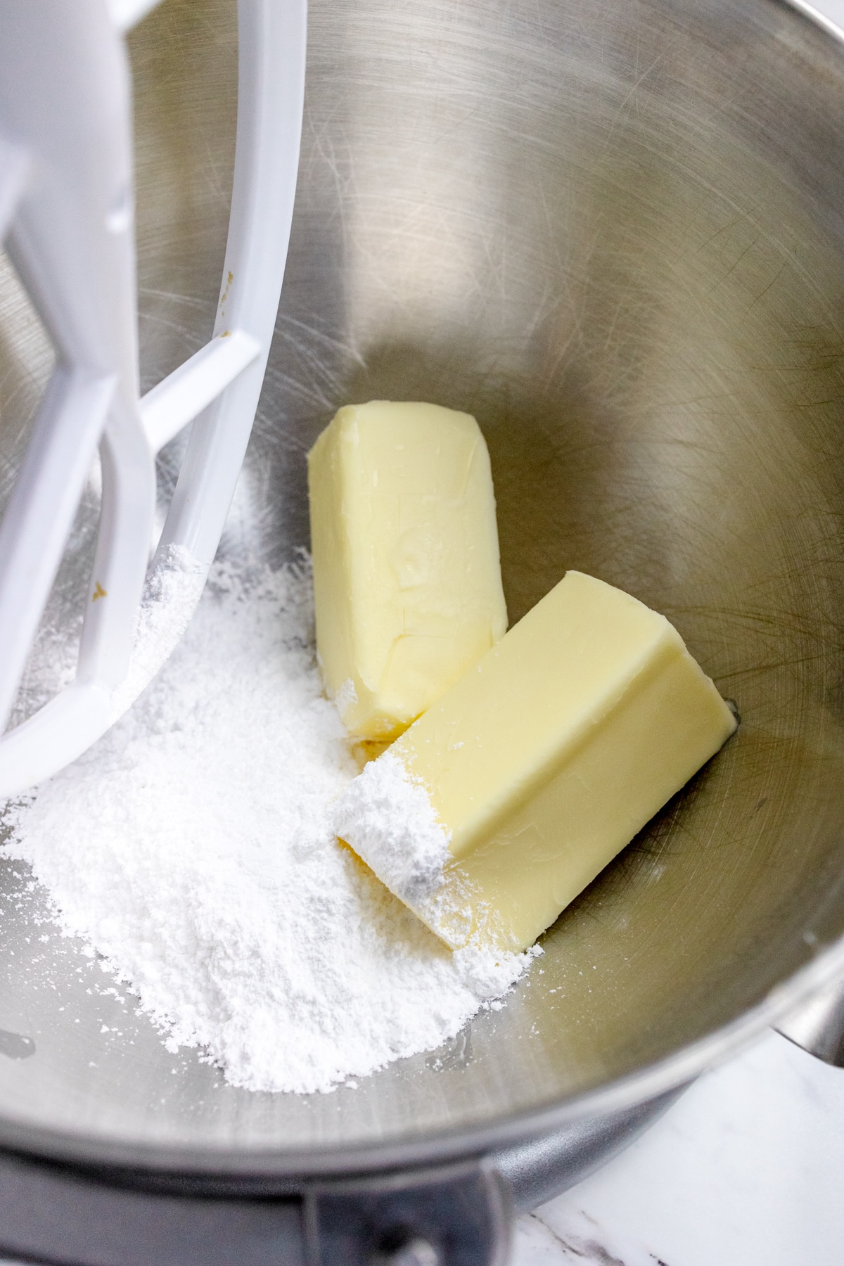 Close up of the bowl of a stand mixer with a paddle attachment, with butter and powdered sugar in the bowl.