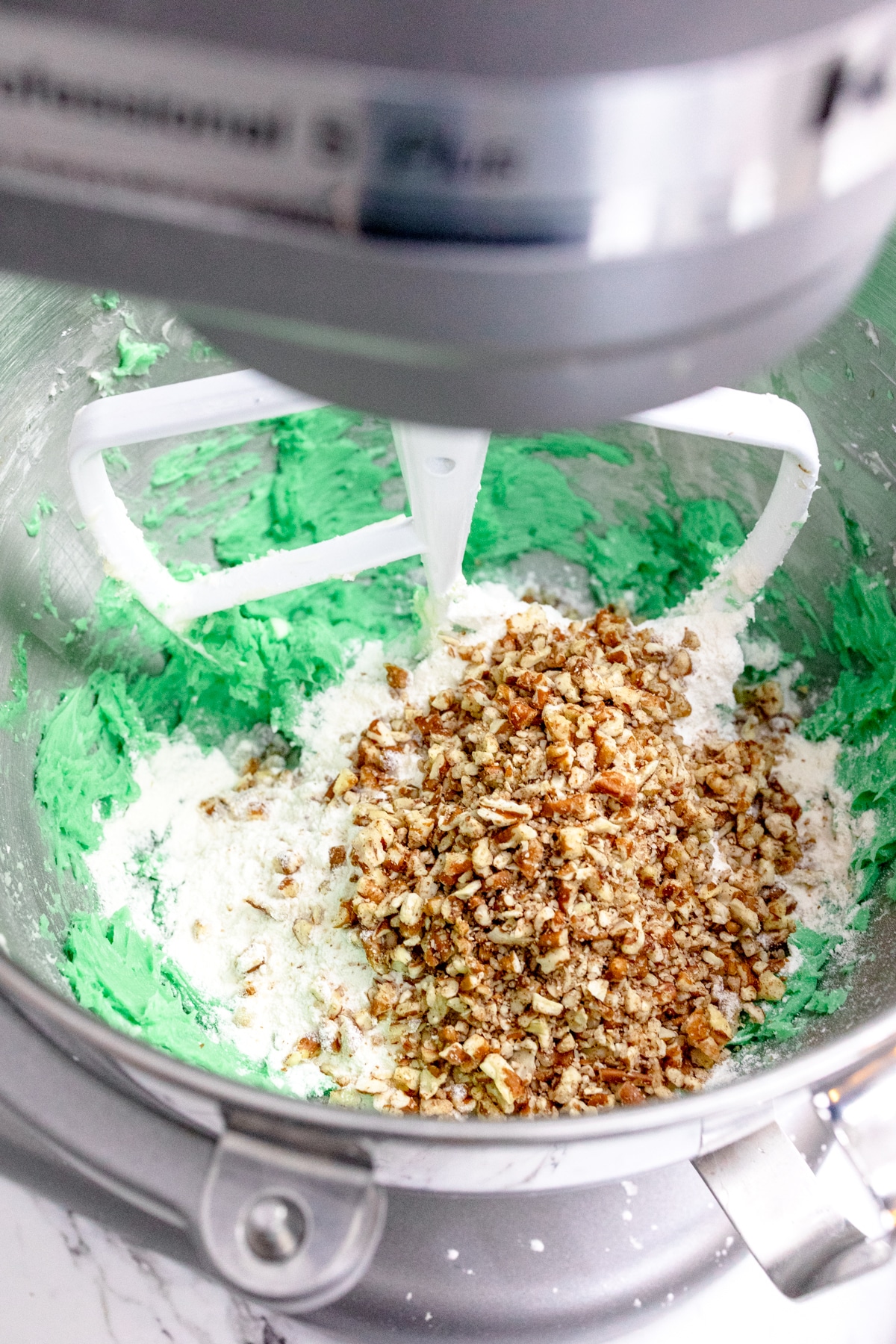 Close up of the bowl of a stand mixer with green cookie dough in it with chopped pecans that have been added on top to mix in.