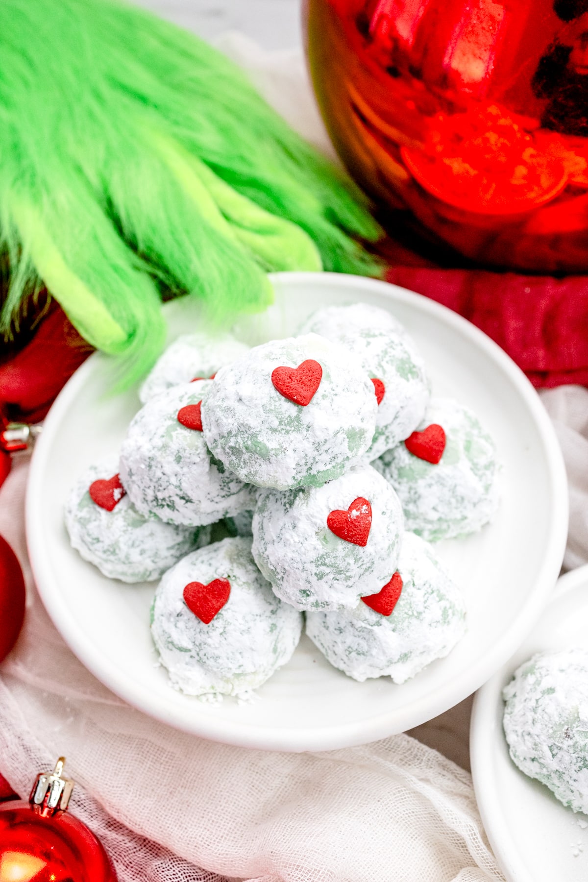 Close up of Grinch Snowball Cookies on a white plate, with Christmas decorations around them, and a furry green hand reaching for them.
