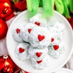 Close up of Grinch Snowball Cookies on a white plate, with Christmas decorations around them, and a furry green hand reaching for them.
