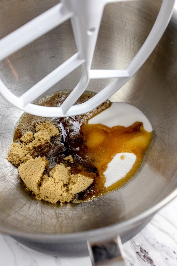 Close up of the bowl of a stand mixer with brown butter, white sugar, and brown sugar ready to be creamed together with a paddle attachment.