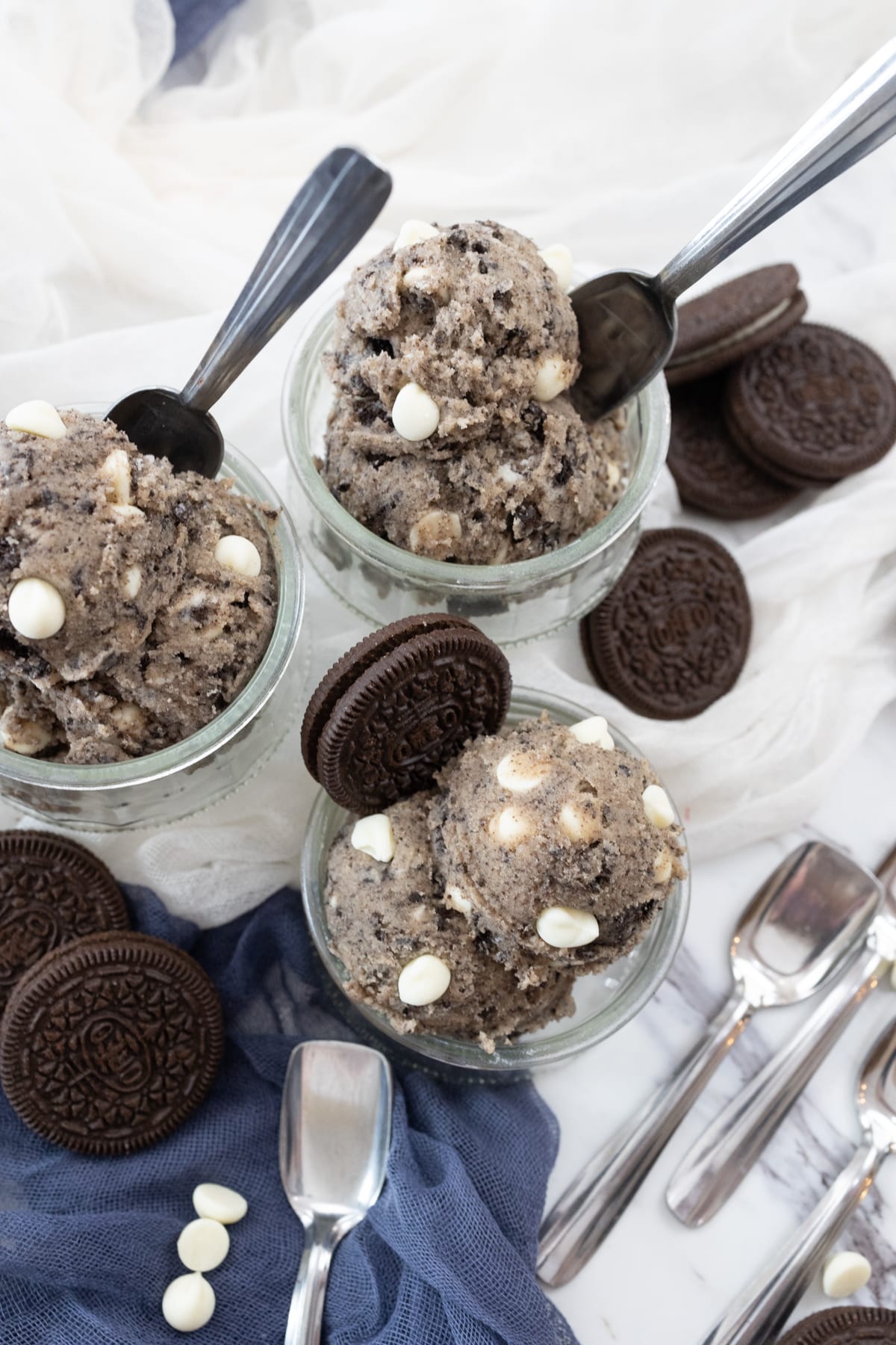 Top view of small glass bowls containing Edible Cookie Dough that has a spoon and an oreo cookie sticking out of it like an ice cream wafer, on a surface which has white and blue fabric on it for decoration.