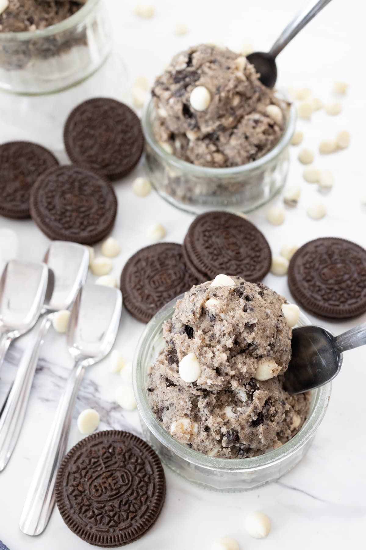 Top view of small glass bowls containing Edible Cookie Dough that has a spoon and an oreo cookie sticking out of it like an ice cream wafer, on a white surface.