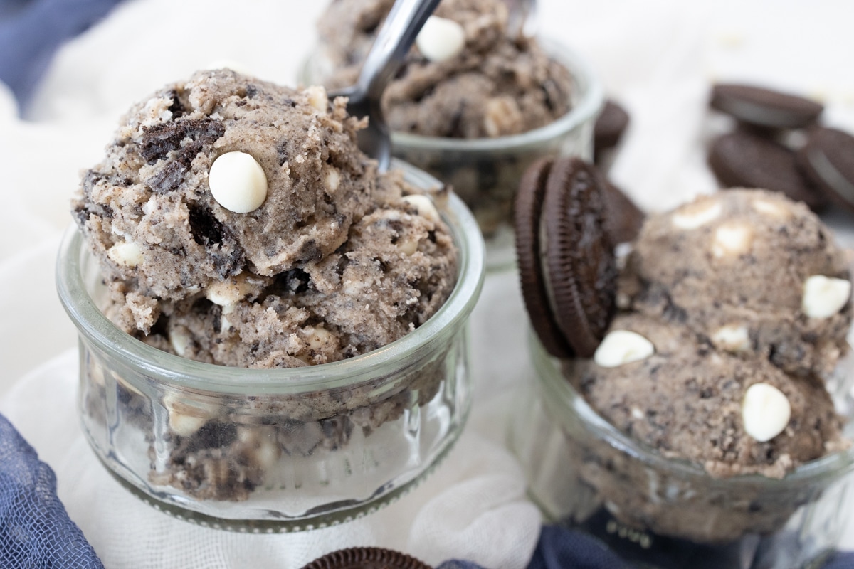 Top view of small glass bowls containing Edible Cookie Dough that has a spoon and an oreo cookie sticking out of it like an ice cream wafer, on a surface which has white and blue fabric on it for decoration.