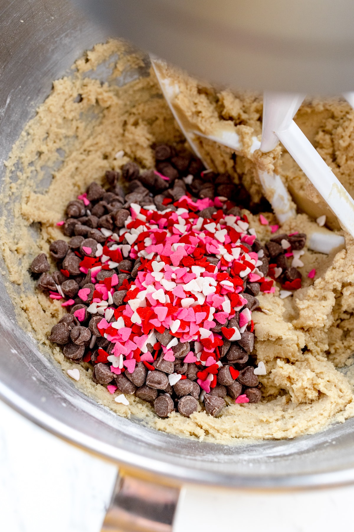 Close up of the bowl of a stand mixer with cookie dough in it with chocolate chips and sprinkles that have been added on top to mix in.