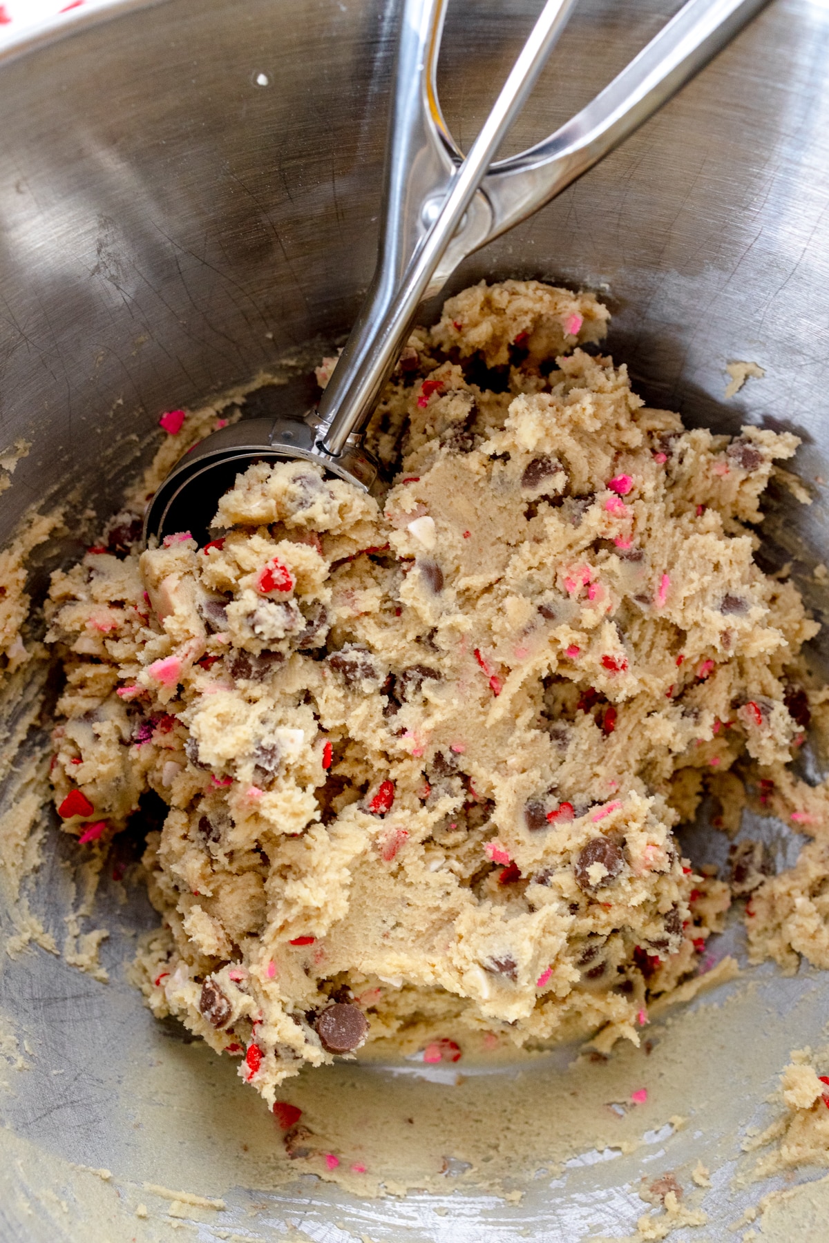 Close up of the bowl of a stand mixer with cookie dough in it and a cookie scoop scooping some out.