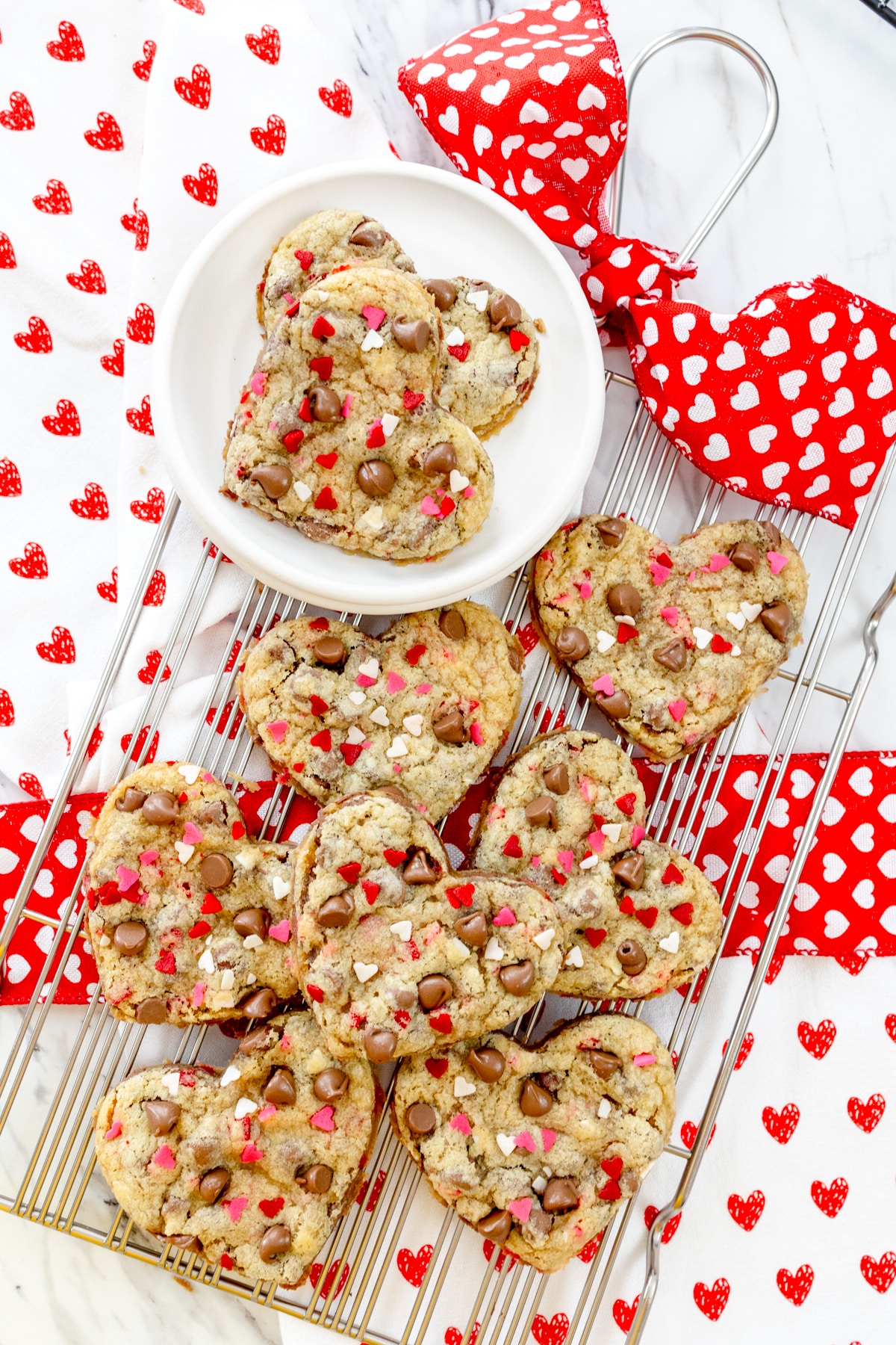 Top view of two heart shaped cookies on a white plate, which is on a wire rack that has other heart shaped cookies on it, and there are love heart decorations around the cookies.