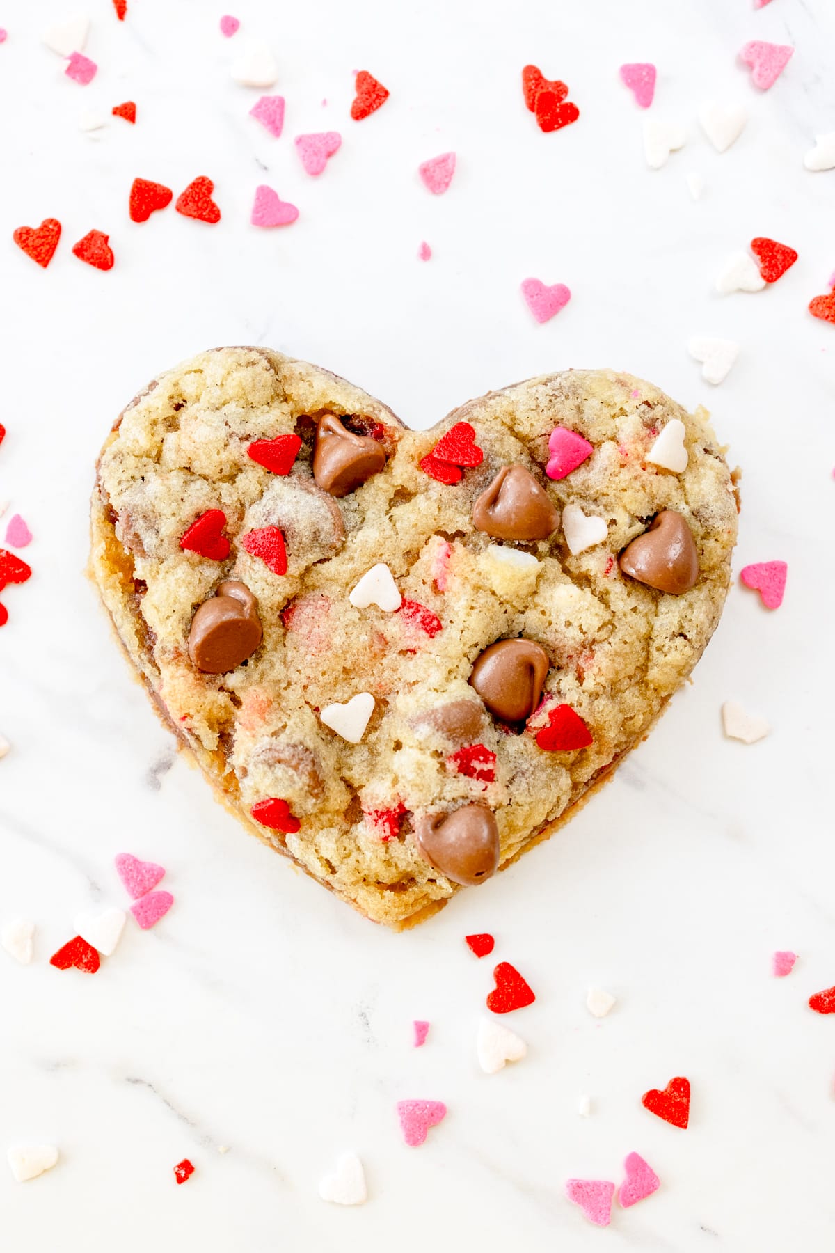 Top view of a heart shaped cookie on a white surface, and there are love heart decorations around the cookie.