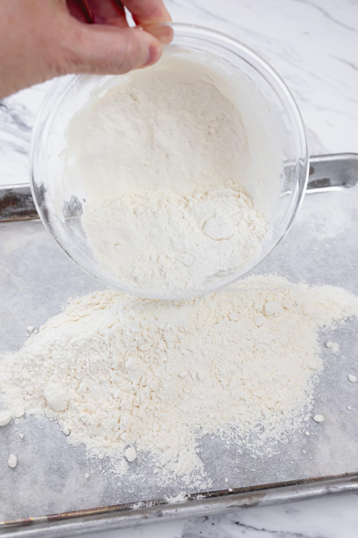 Close up view of flour in a microwavable bowl being poured onto a parchment-lined baking sheet.