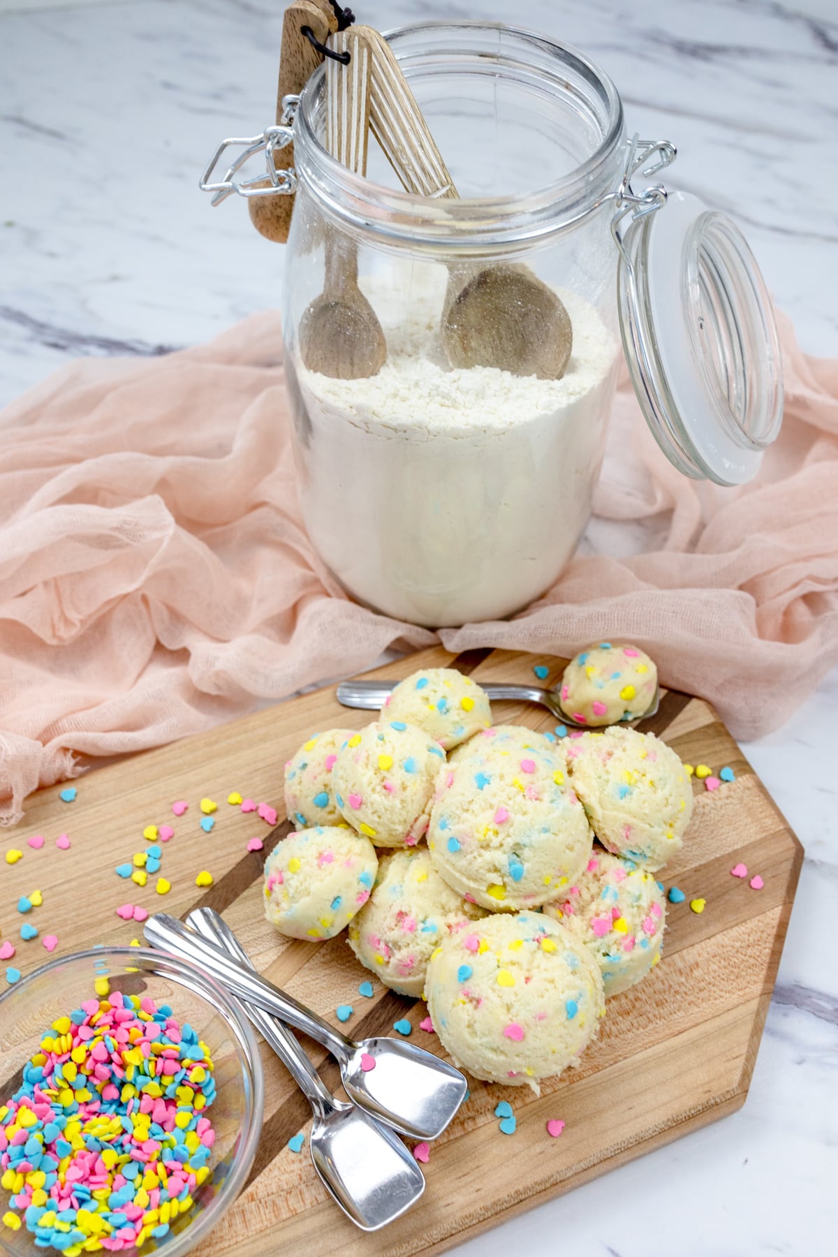 Close up view of edible cookie dough balls on the countertop, beside a storage jar of heat treated flour and a small bowl full of sprinkles.