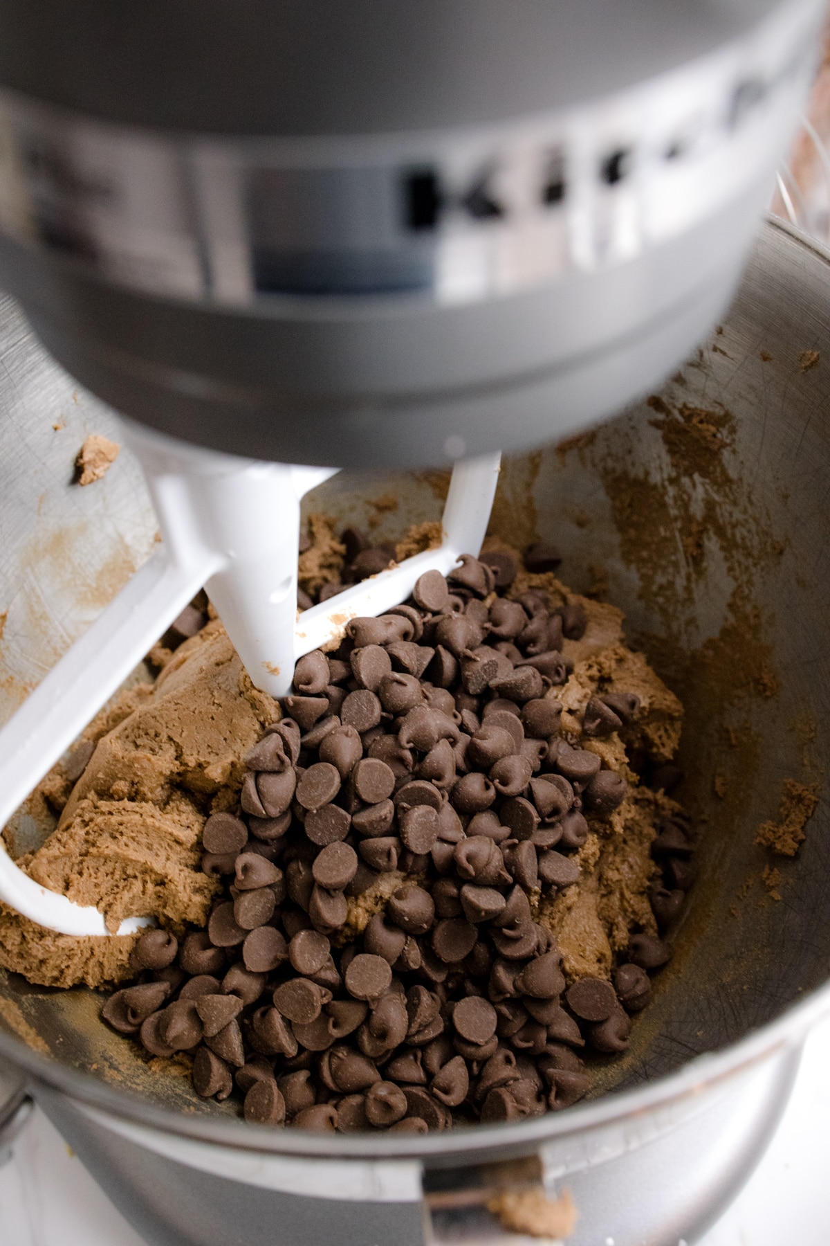 Close up of the bowl of a stand mixer with cookie dough in it with chocolate chips that have been added on top to mix in.