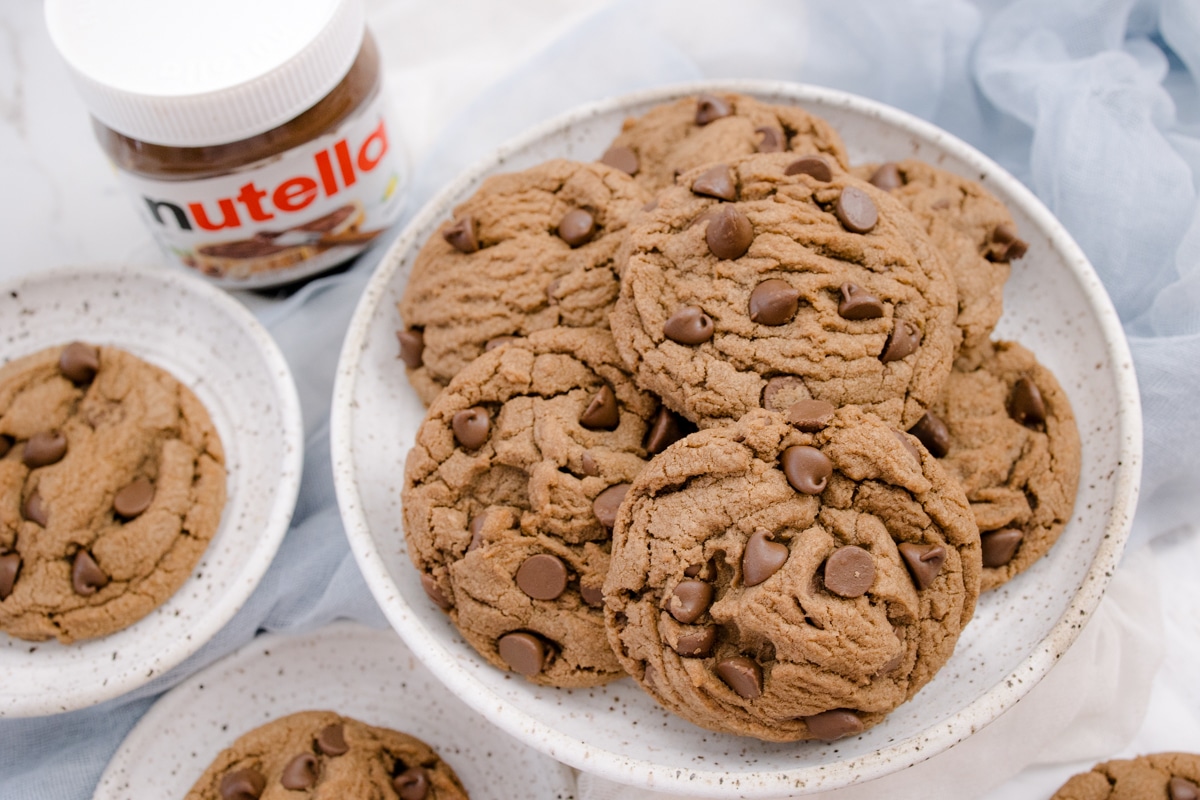 Angled view of Nutella Cookies on several white plates, on a white surface, with a jar of Nutella in the background.