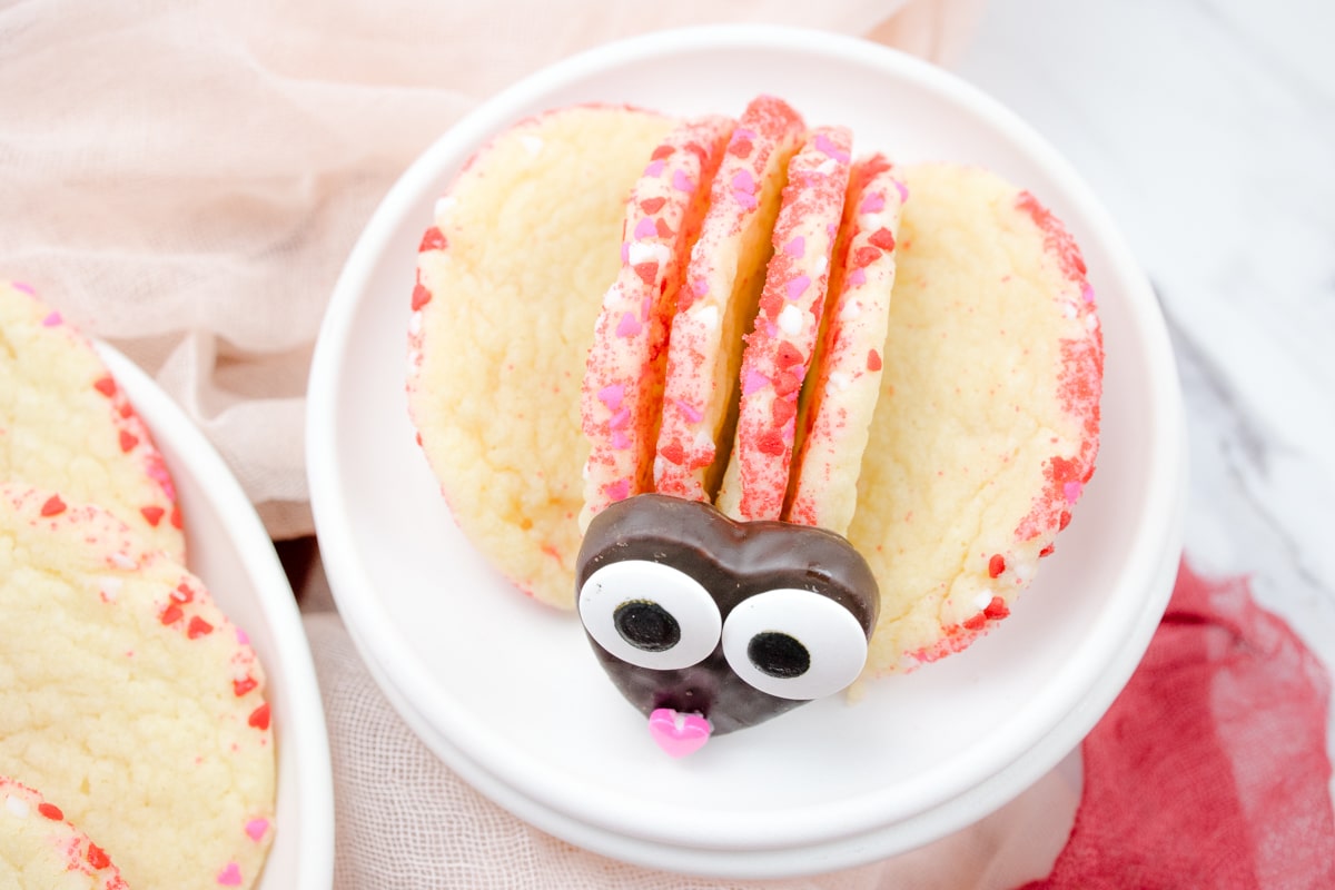 Close up view of a plate with Slice and Bake Sugar Cookies on it, arranged to look like a Love Bug with a body and wings made out of the cookies, and a head made from a chocolate heart with googly eyes and a pink heart sprinkle for a nose.