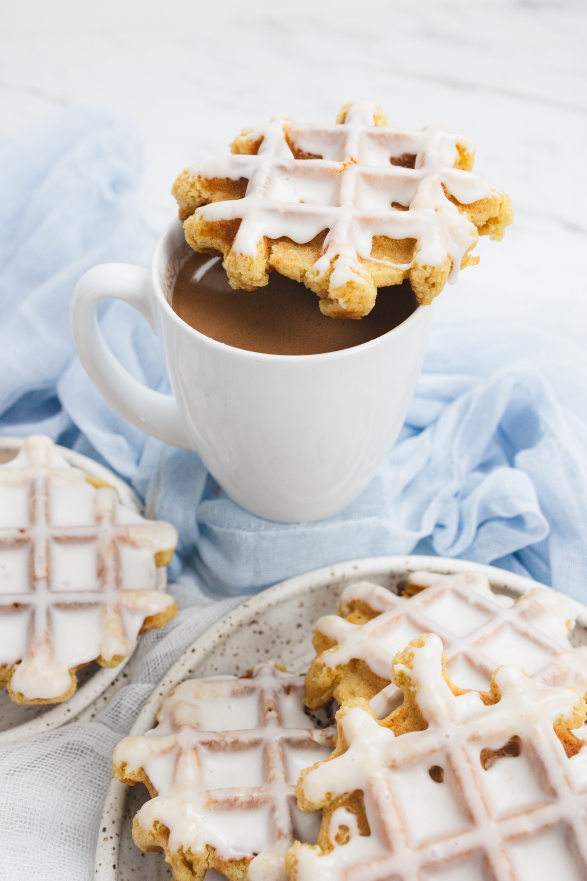 Close up of waffle cookies on a white surface, with a white mug of coffee in the back with a waffle cookie balanced on top of the mug.