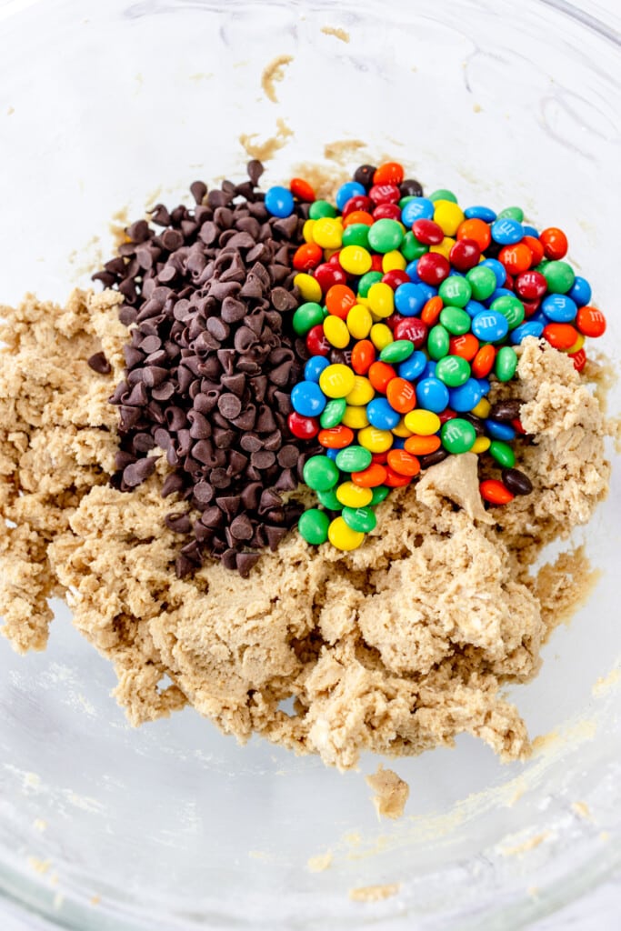 Close up view of a large mixing bowl with m&m's and chocolate chips on the cookie dough, ready to be mixed in.