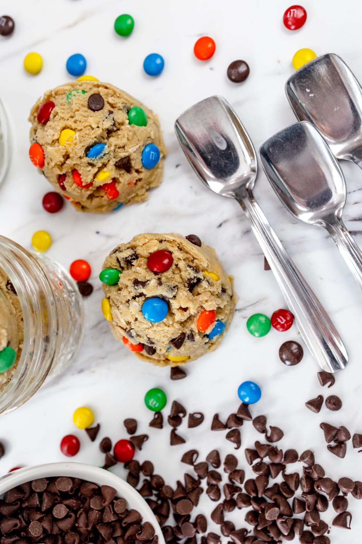Close up view of Monster Cookie Dough in serving bowls on a white counter with a bowl of chocolate chips and silver ice cream spoons around them.