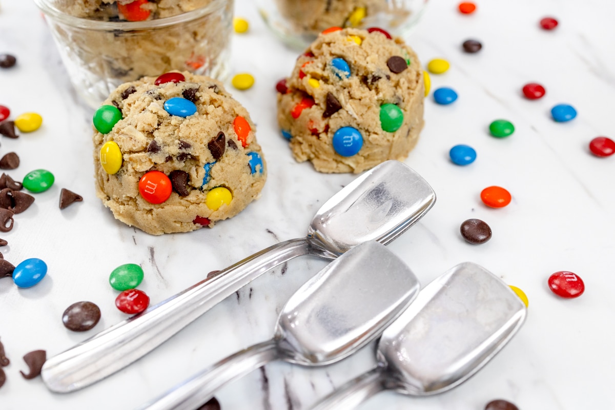 Close up view of Monster Cookie Dough in serving bowls on a white counter with a bowl of chocolate chips and silver ice cream spoons around them.