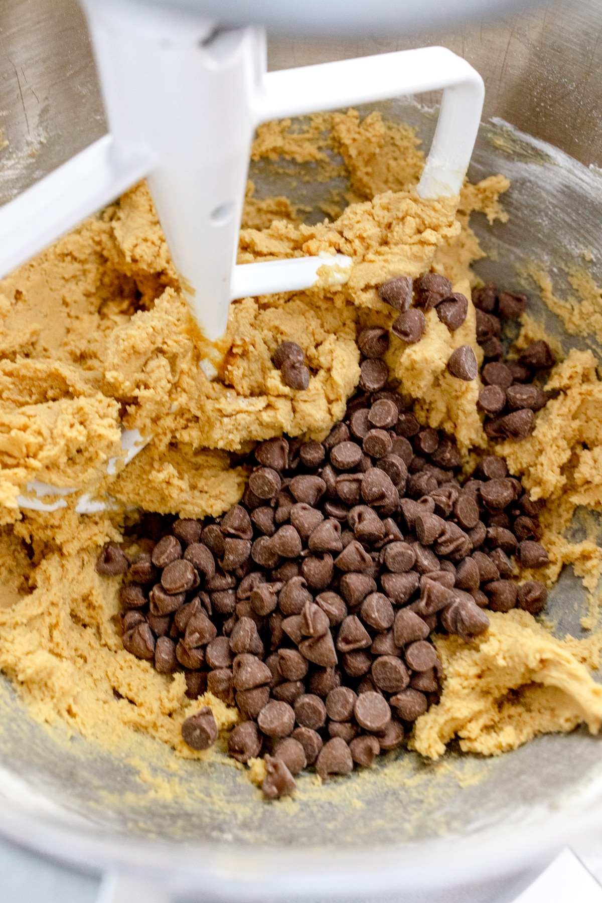 Close up of the bowl of a stand mixer with cookie dough in it with chocolate chips that have been added on top to mix in.