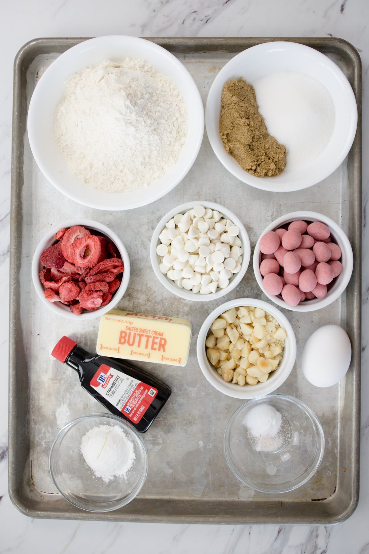 Top view of ingredients needed to make Strawberry White Chocolate Macadamia Nut Cookies in small bowls on a baking tray.