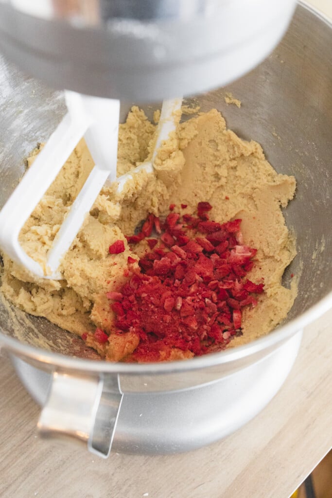 Close up of the bowl of a stand mixer with cookie dough in it with freeze-dried strawberries that have been added on top to mix in.