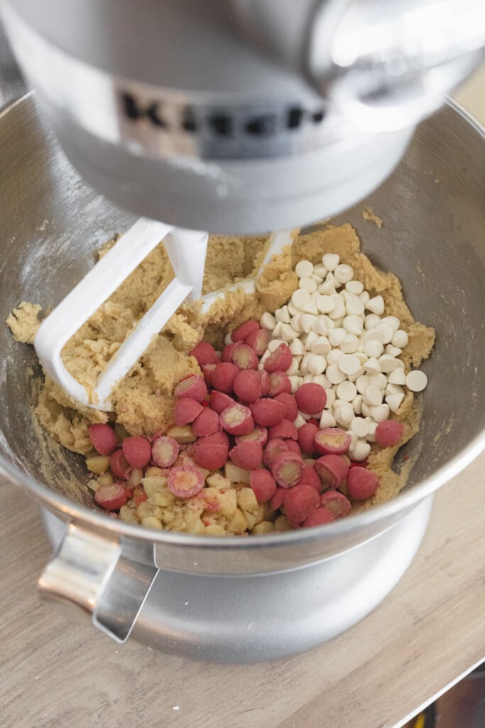 Close up of the bowl of a stand mixer with cookie dough in it with strawberry chocolate macadamia nuts and white chocolate chips that have been added on top to mix in.