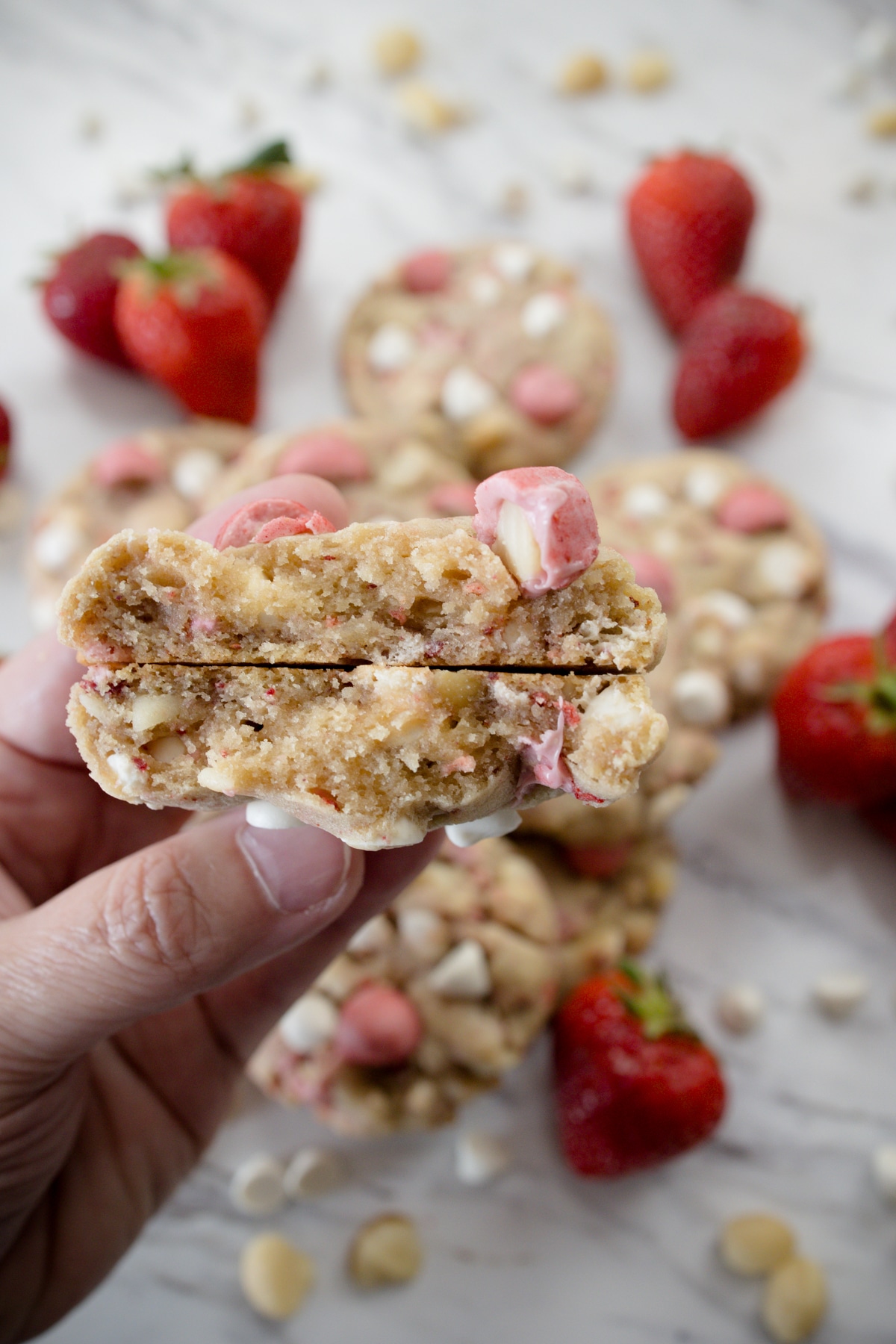 Close up view of a hand holding a Strawberry White Chocolate Macadamia Nut Cookie which has been broken in hald to reveal the middle, in front of a white surface with other cookies and decorations on it.