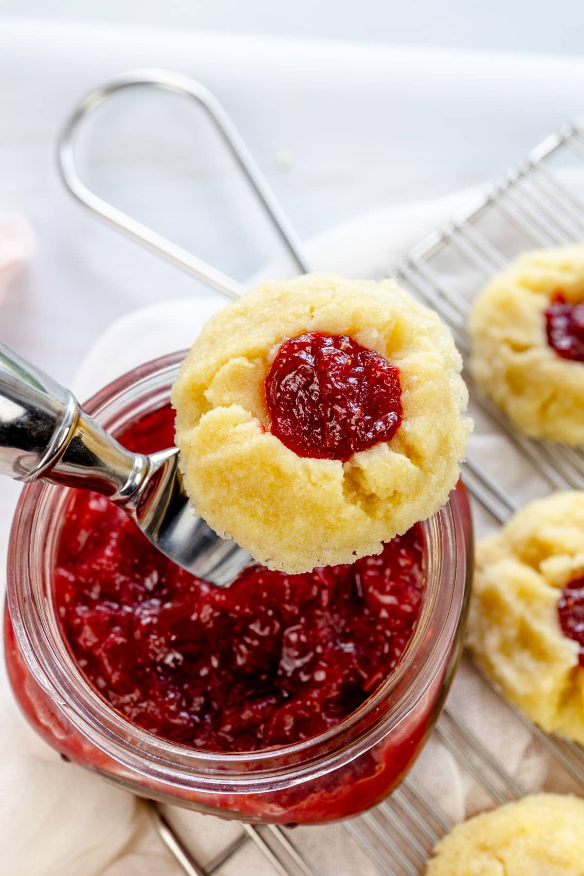 Close up image of a Fig Jam Cookie resting on top of a jar of fig jam with a knife in it, next to a wire rack with fig jam cookies on it.